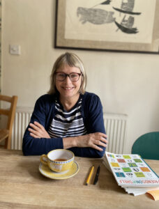 A woman with short blonde hair and spectacles sits at a kitchen table, with a cup of tea and a magazine in front of her